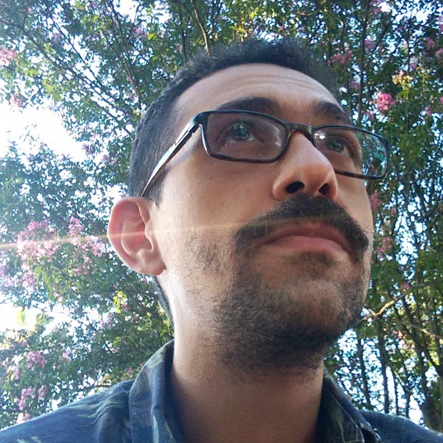 Imagem de homem moreno, cabelos pretos e curtos, bigode e barba curta, com fundo arborizado e florido